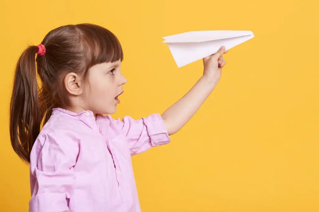 Side view of little cute female kid posing with white paper plane in hands, charming concentrated girl wearing rose shirt, having dark hair, female child being fond of origami. Childhood concept.